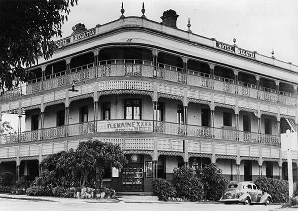 This is a black and white photograph of the Regatta Hotel circa 1940. From a corner view it shows the 3-storey hotel with its balconies, ornate railings and fine pillars stretching the length of each side on the second and third levels. A banner of the corner entrance of the second storey balcony reads 'Castlemaine XXXX Beer'.