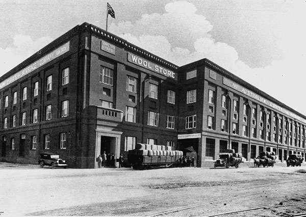 A black and white photograph of the Australian Estates No.1 Store in 1928 shows a very large 3-storey brick building. Both horse-drawn wagons and automotive delivery trucks loaded with wool bales are parked outside on the dirt road.