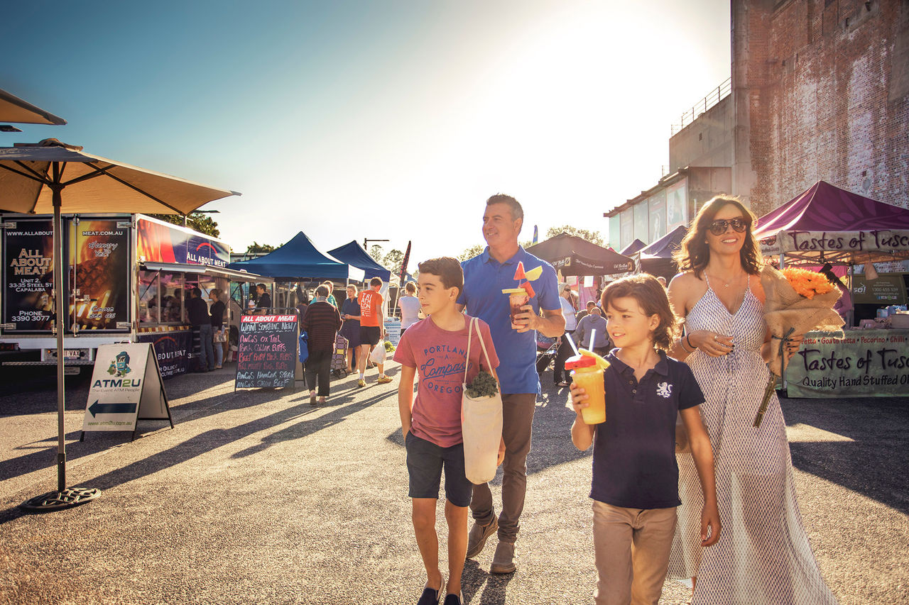 A family of four walking through the Jan Powers Markets at Brisbane Powerhouse. Each person is carrying a different market item.