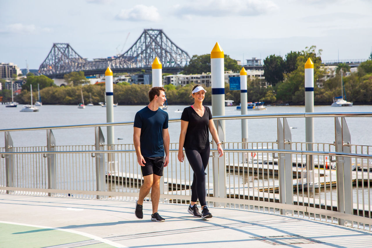 Man and woman walking along the City Botanic Gardens riverwalk, alongside the Brisbane River, with the Story Bridge in the background.