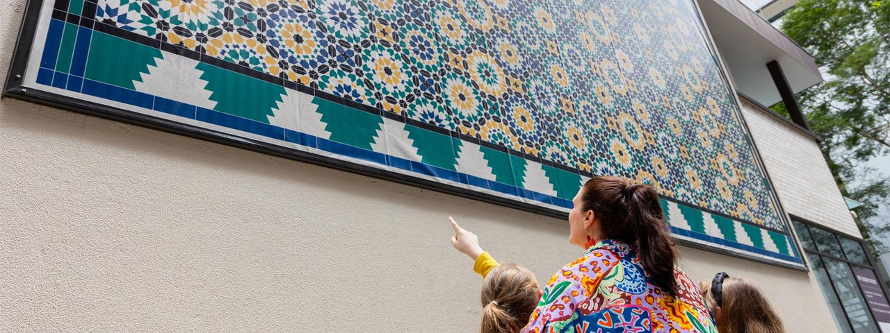 A woman and two children look up at the colourful Edison Lane banner, featuring intricate geometric patterns on a building's exterior wall.