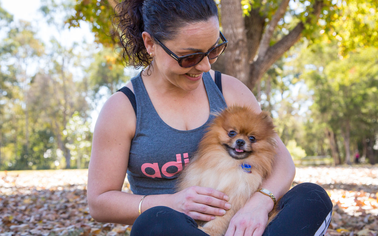 Female in active wear sitting cross-legged in a shady Council dog park with a Pomeranian dog on her lap. Dog is wearing a blue registration tag.
