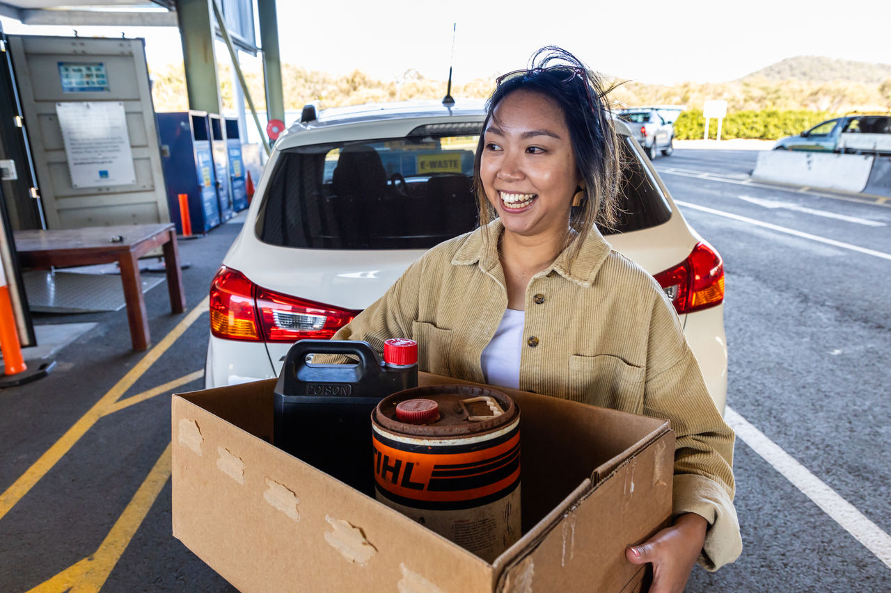 Woman carrying a cardboard box with oil and mower fuel for a hazardous waste drop-off at Chandler Resource Recovery Centre. Her white hatchback vehicle is in the background.