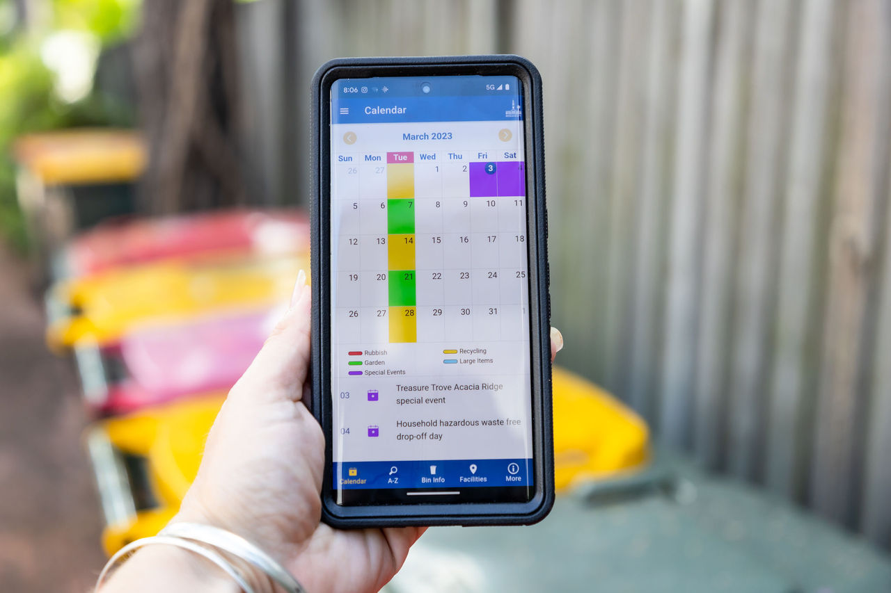Woman holding the Brisbane Bin and Recycling app in her left hand. Bins (general and recycling) for multi-unit dwelling complex in background.