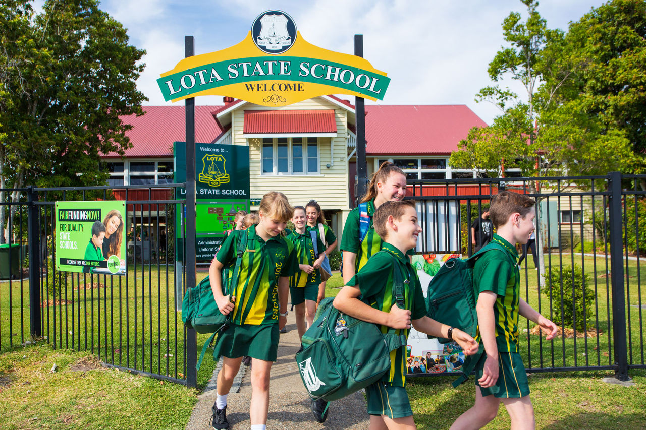 Group of older primary school students in uninform leaving Lota State School through school gate. School in background.