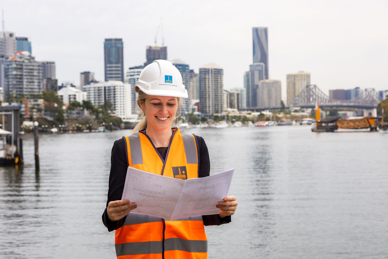 Female engineer looking at plans onsite with Brisbane CBD and Brisbae River in background.