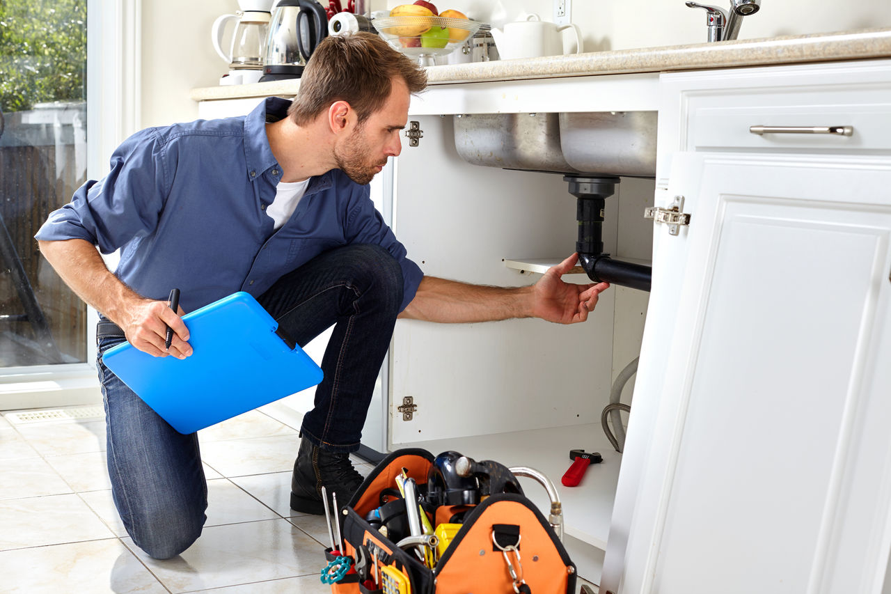 Plumber crouching down to inspect under sink pipes in a modern, white, domestic kitchen.