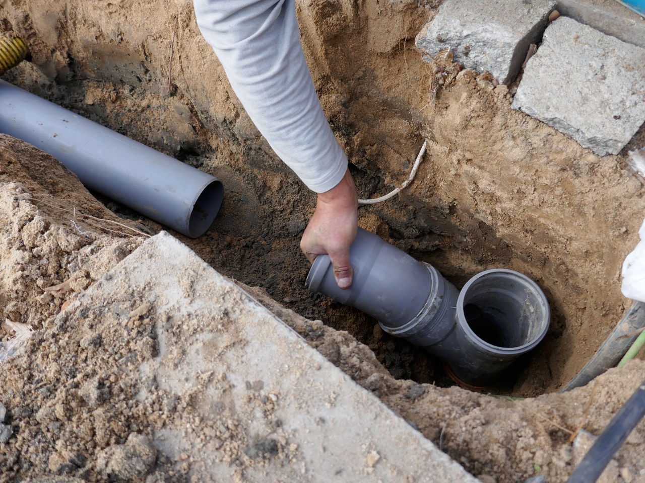 Close up of a man's hand placing a grey sewer pipe in a trench.