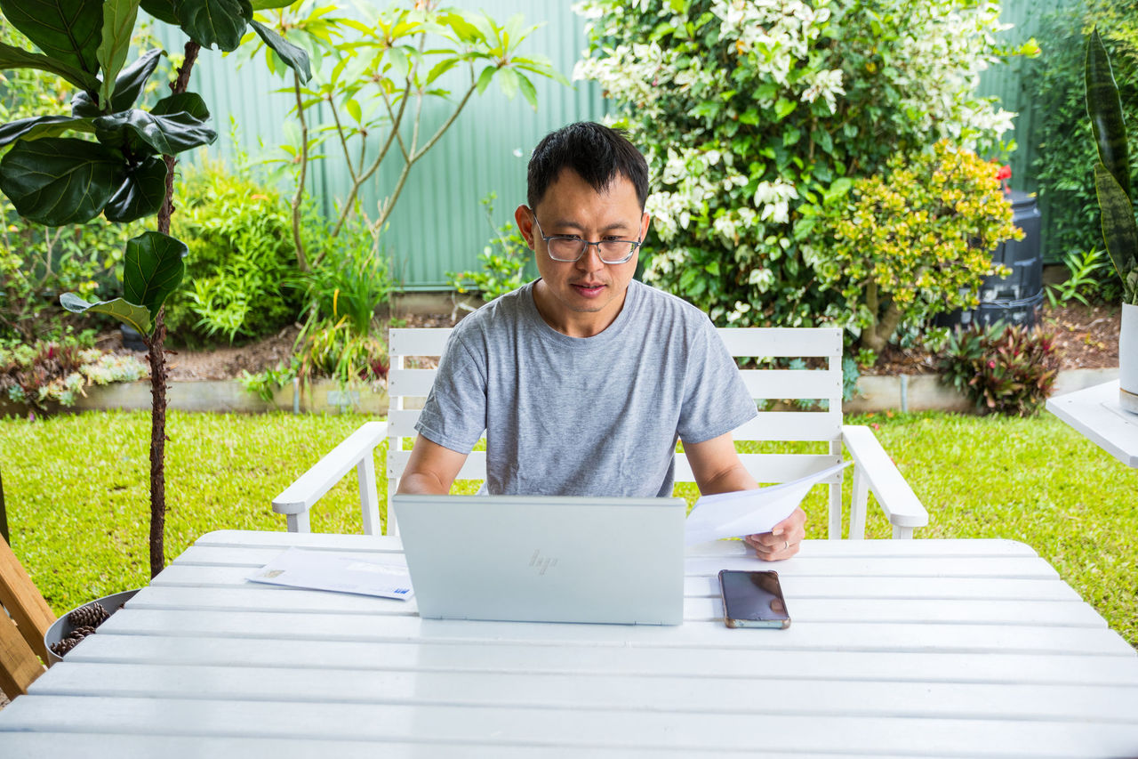 Man using a laptop at an outdoors table with a piece of paper in his left hand and mobile phone on table.