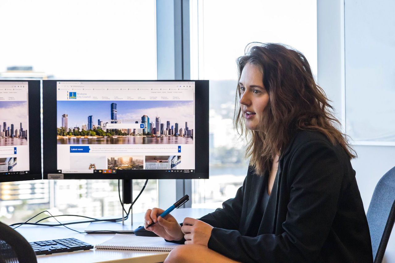 Young professional in a business setting at a desk and talking with someone (off screen).