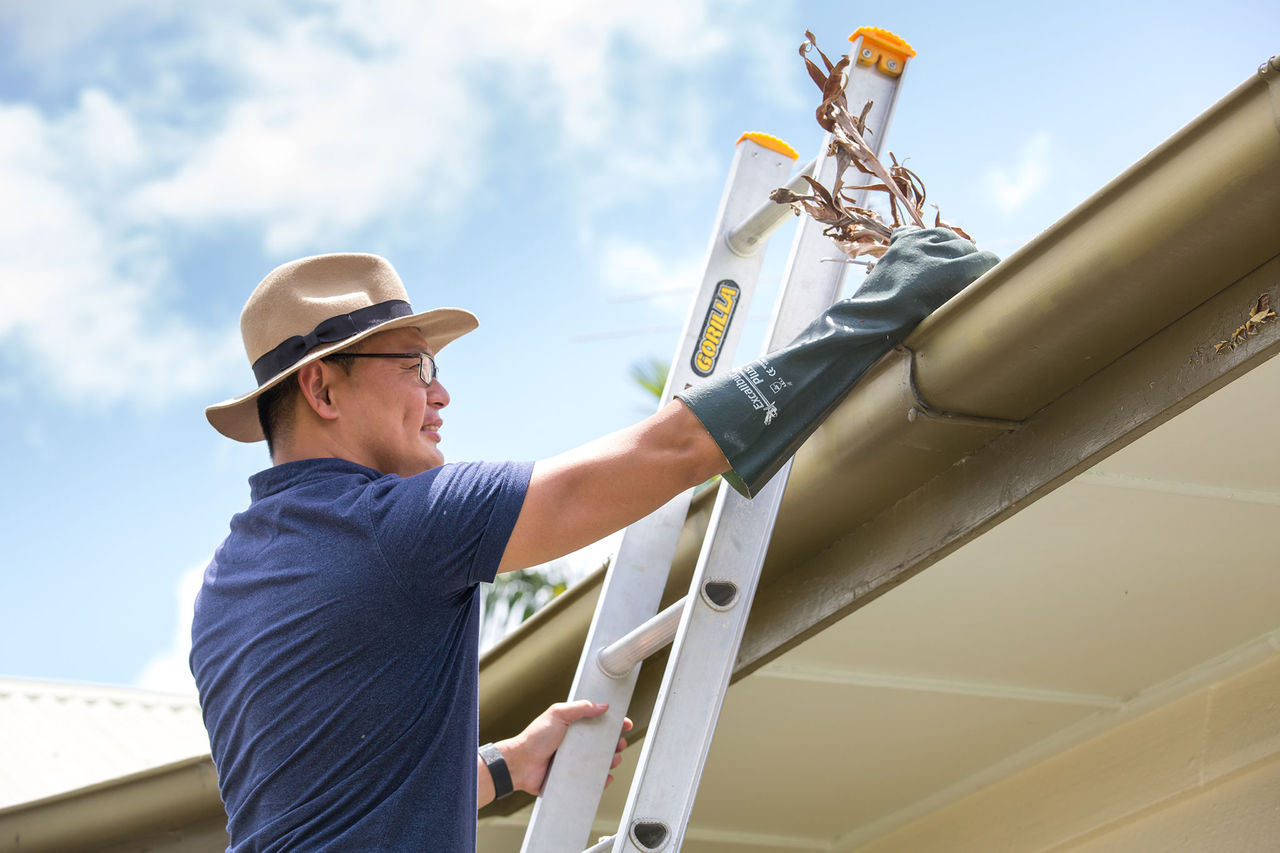 Person standing up on a ladder getting leaves and branches out of roof guttering.