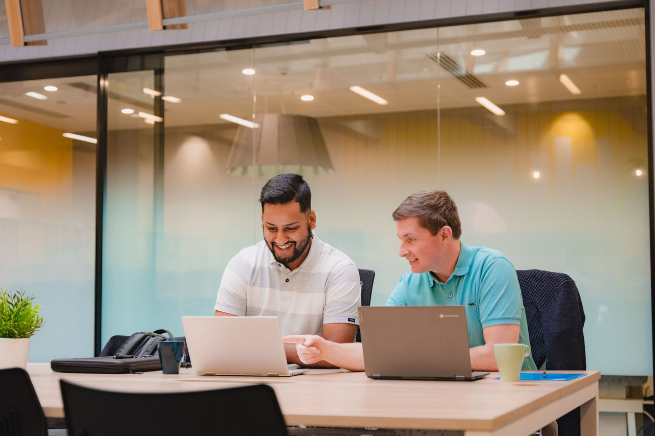 Two men working on laptops and talking with one another in an open plan office.