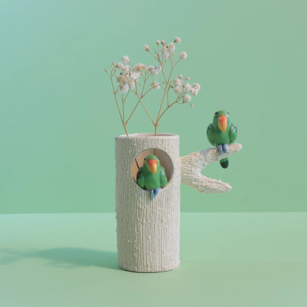 This image shows a ceramic white vase in the shape of a tree trunk. It holds small white flowers and features 2 colourful ceramic parakeets.