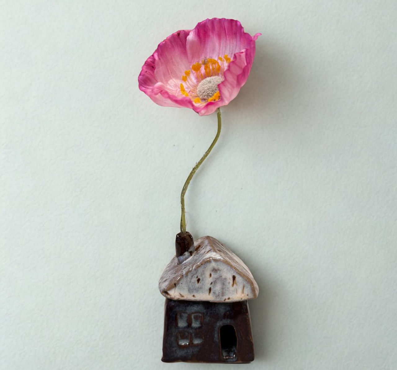 This is an image of a ceramic house artwork by D H Designs. There is a pink poppy artificial flower extending from the chimney.