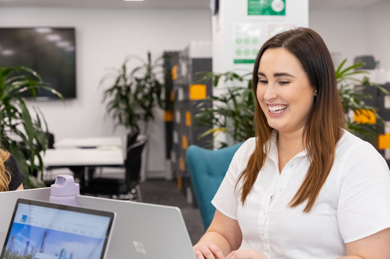 Smiling woman working on a laptop in a modern, open-plan office.