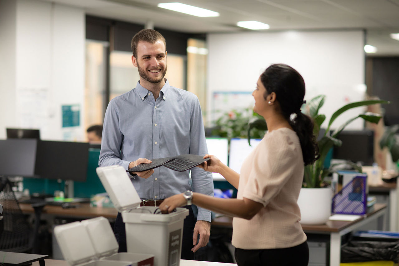 Two people in an office standing around a desk placing items for recycling into a bin.