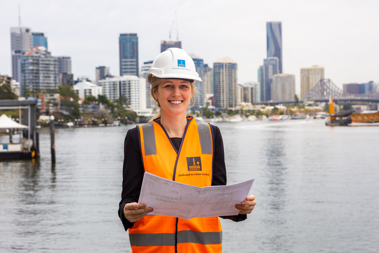 Female engineer on a development site at Kangaroo Point with A3 plans open in hands. Site is on Brisbane River, with Kangaroo Point buildings, Story Bridge and Brisbane CBD in background.