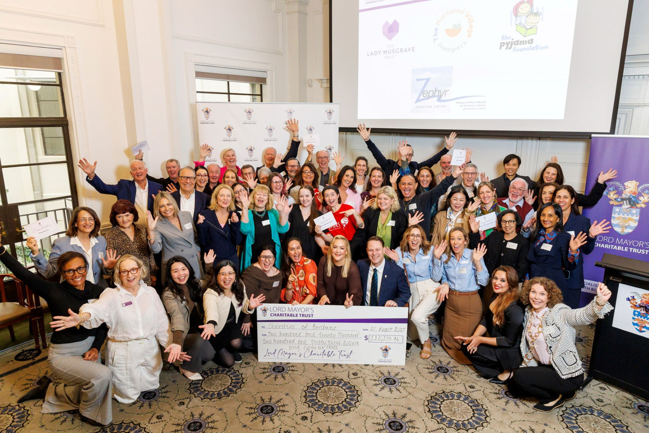 This image shows a large group of people smiling with their hands up. At the front, a few people hold a large cheque with the “Lord Mayor’s Charitable Trust” written on it.