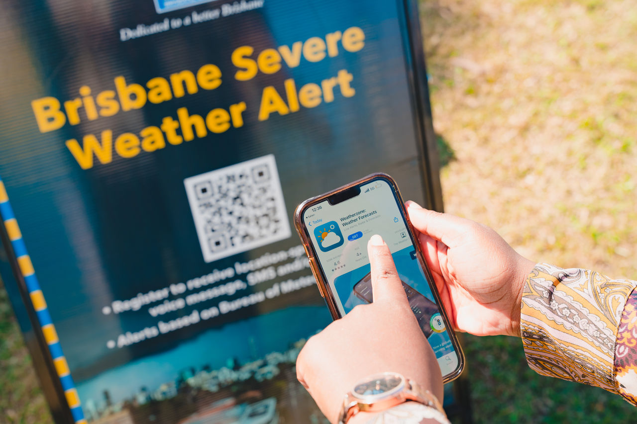 Close-up of a woman's hand at a fair standing in front of a Brisbane Severe Weather Alert advertising A-frame. and pointing to Weatherzone app on smartphone.
