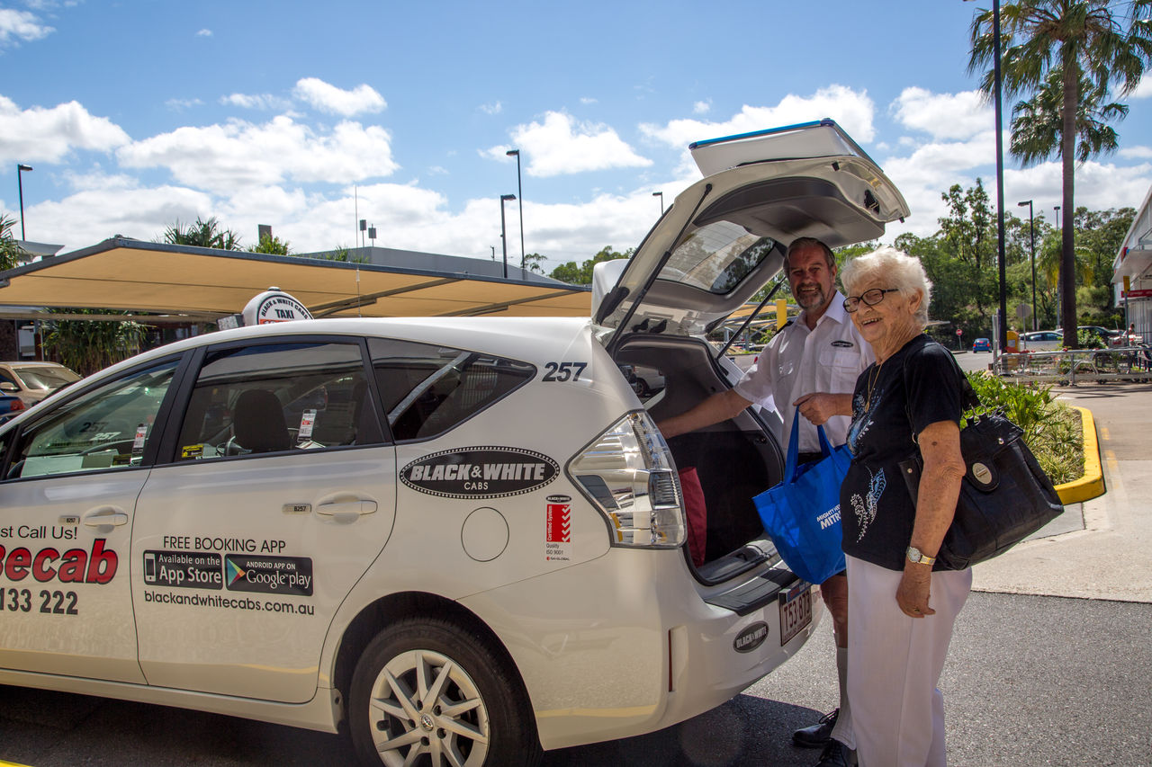 Male Black & White Cab driver helping elderly woman place groceries in boot of sedan.