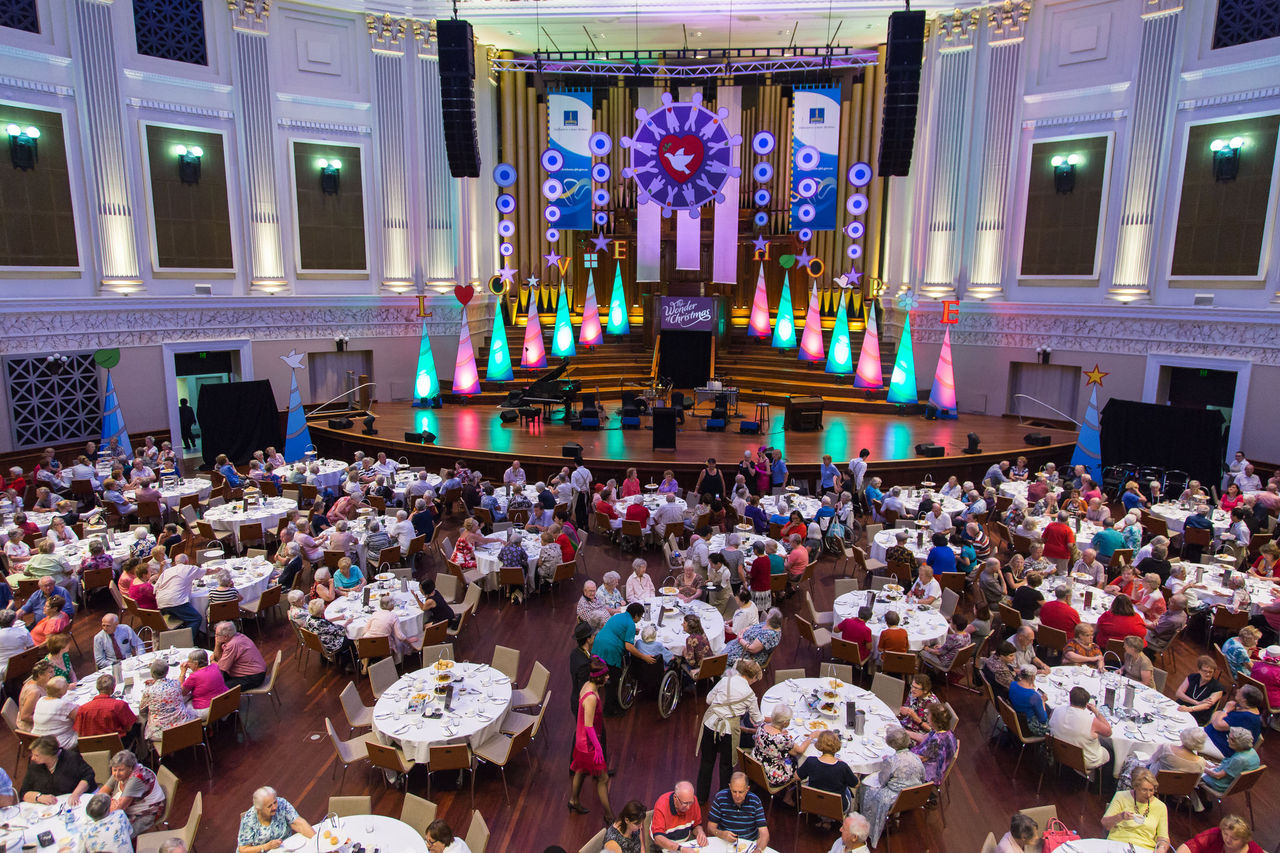 Seniors sit at tables in front of a large stage with Christmas-themed decorations.