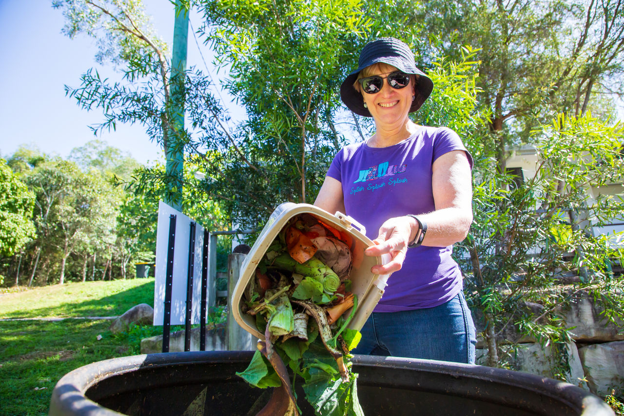 Woman adds green waste to a composting hub with lush trees in the background.