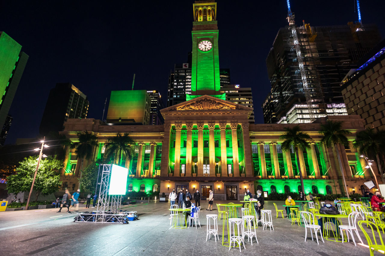 Brisbane City Hall (viewed from King George Square) lit up in green and gold colours. People, screen and tables and chairs in King George Square.