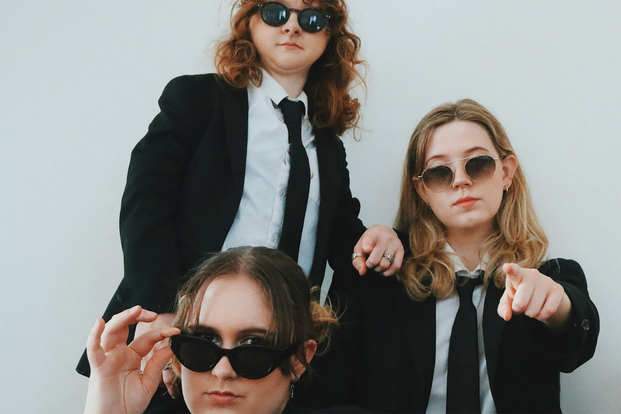 Three band members in Red Hill band - finalists in The QUBE Effect 2026. Photographed against a white wall. All members wearing dark sunglasses.