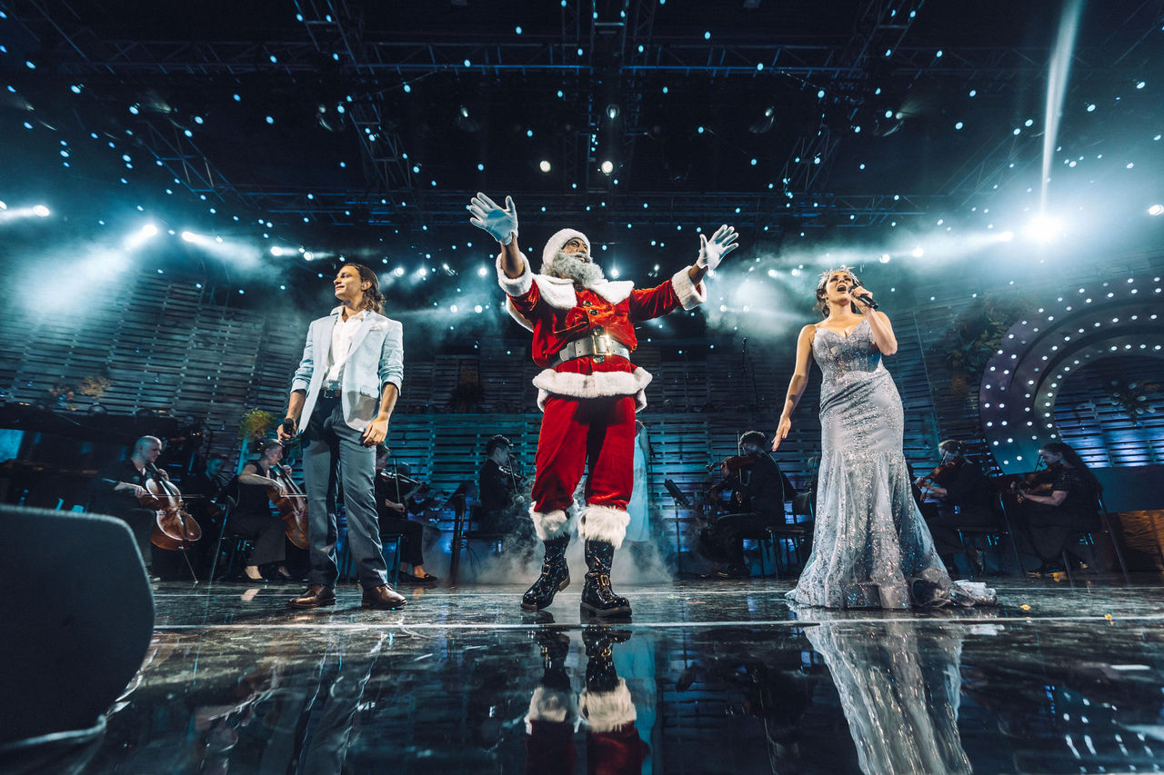 A man dressed as Santa stands on a stage and waves to the crowd. Next to him, a woman sings and a man smiles at the crowd.