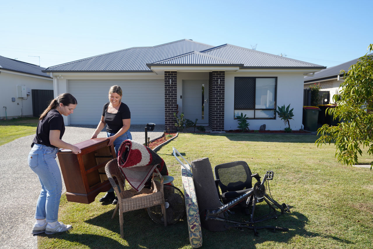 Two women taking large items out to footpath in front of a low-set house for kerbside collection.