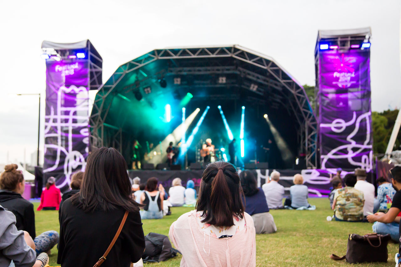 Two girls sitting on the grass, surrounded by crowd at music festival.
