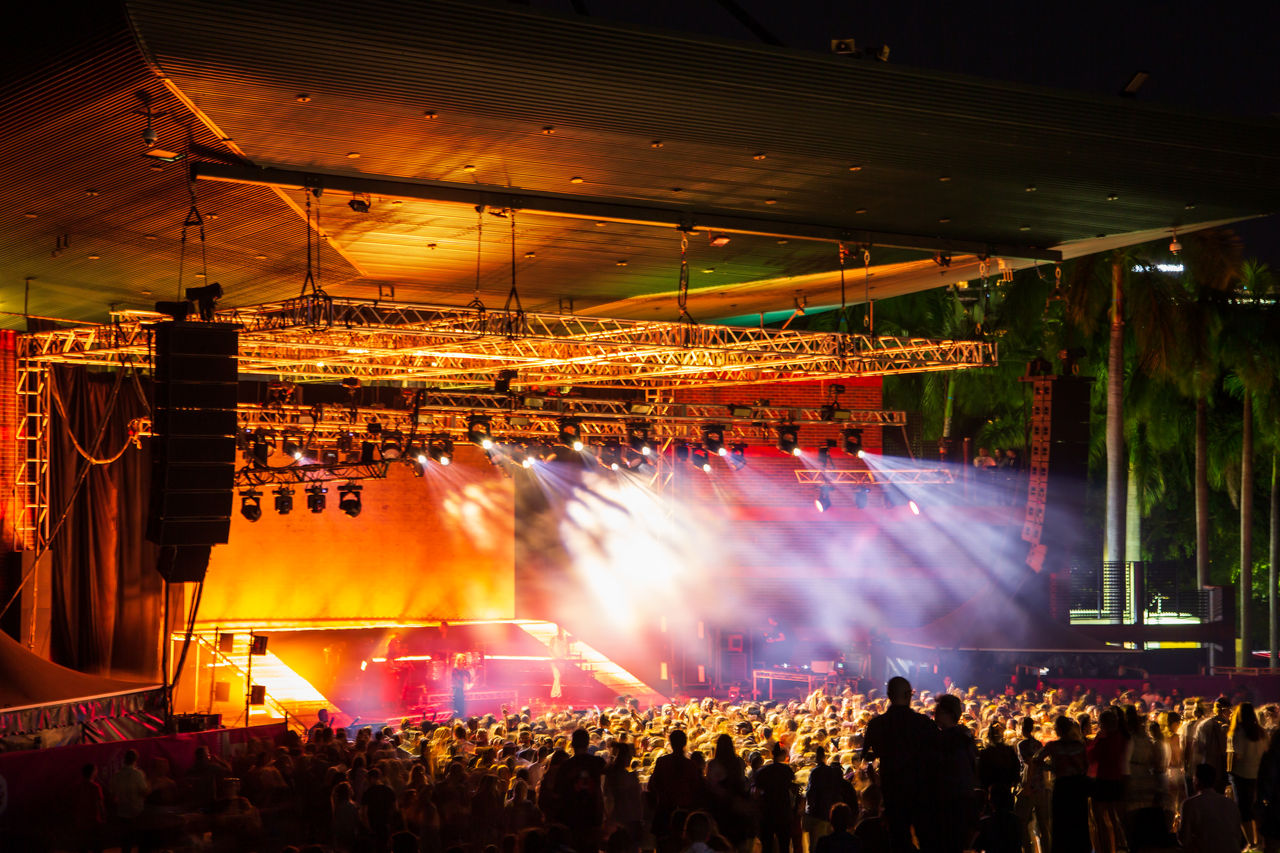 Audience at a Riverstage concert at night. Lighting in orange hues. Image shot from back of venue.
