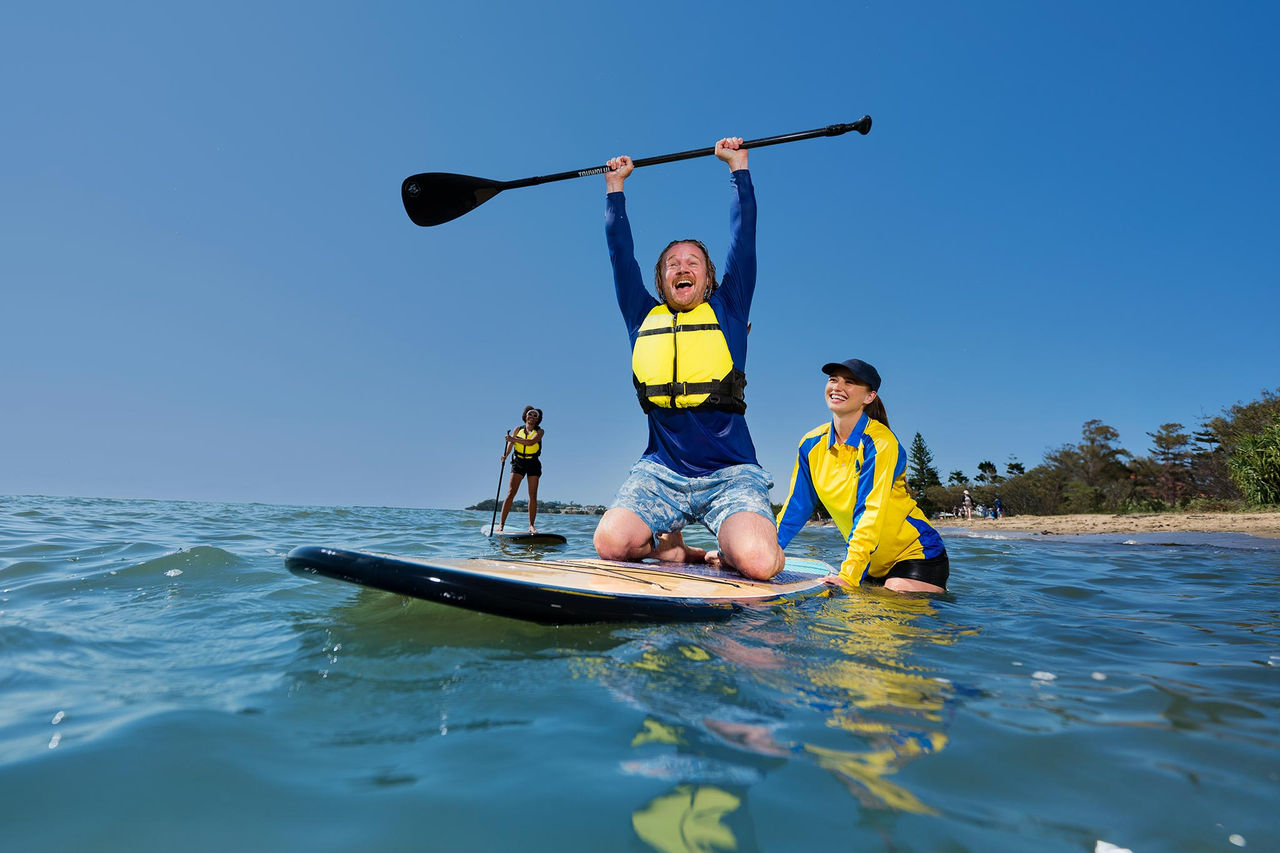 Man on a paddleboard looking happy with oar overhead. Female instructor holding his board.
