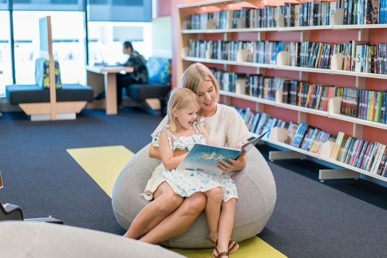 Mother and daughter on a beanbag-style seat at a Council library. Books, library furniture and patron in background.