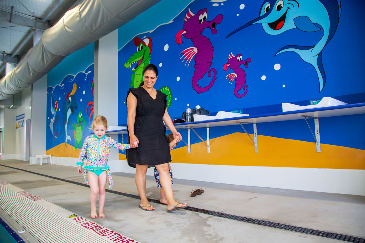 Mum and 2 kids walking into indoor pool at Sandgate Aquatic Centre. Colourful murals on wall behind the subjects.