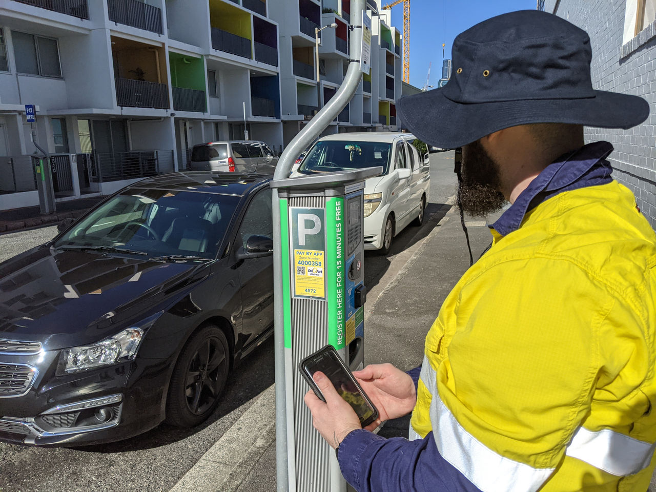 A Council parking inspector wearing personal protective equipment and uniform at a parking meter. Officer has a smartphone in his hand.