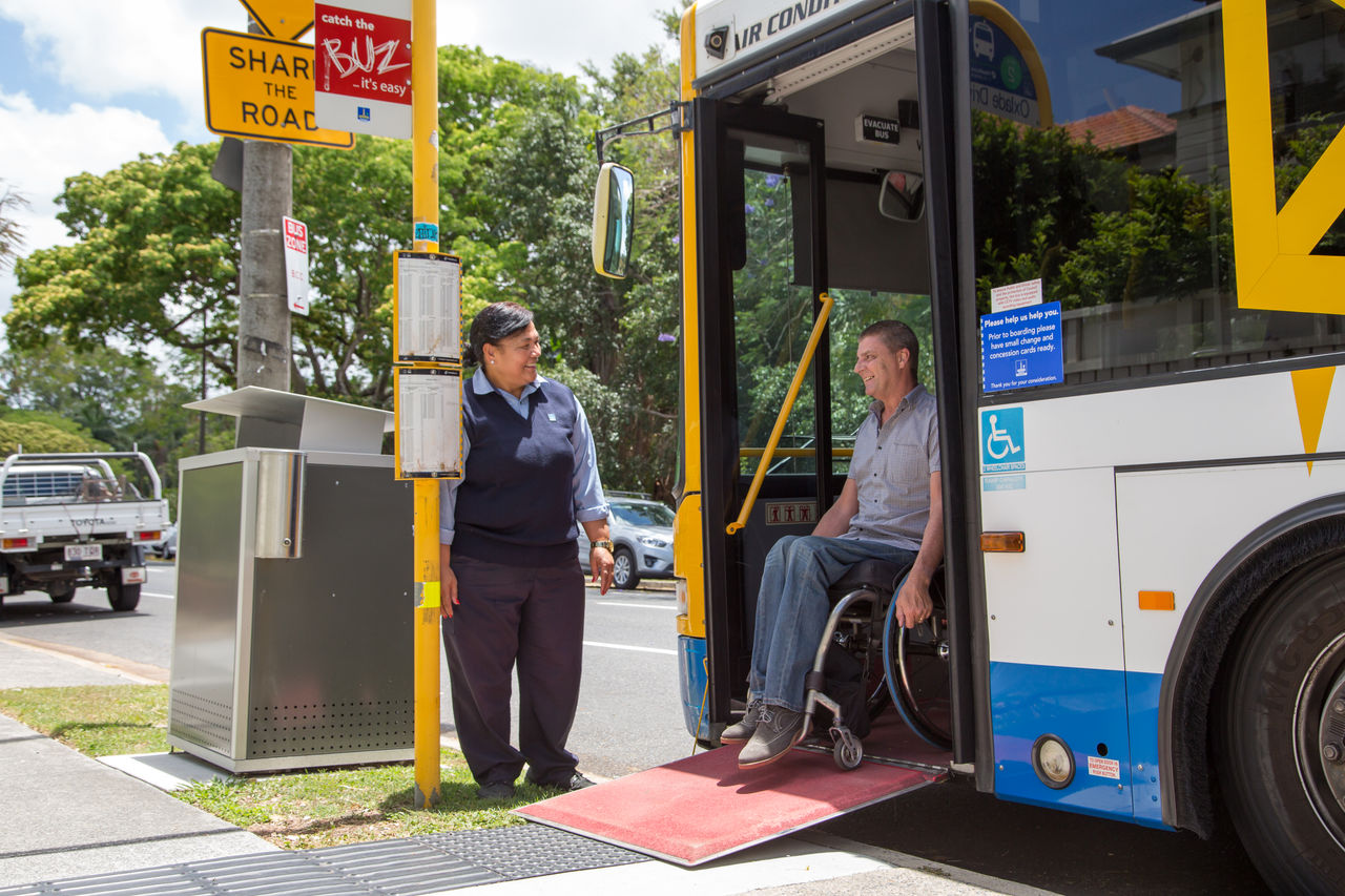 Female Council bus driver helping a male passenger in a wheelchair exit a bus via the front door.