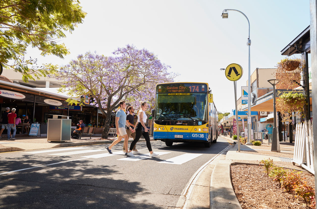A Council bus at a pedestrian crossing in Stones Corner Village. 3 pedestrians midway across the crossing.