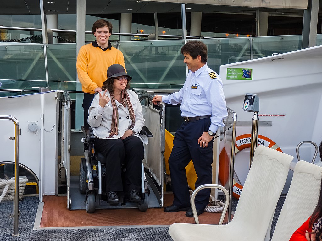 Woman in a wheelchair being wheeled off a CityCat by a young man. Male CityCat officer standing by the CityCat ramp.
