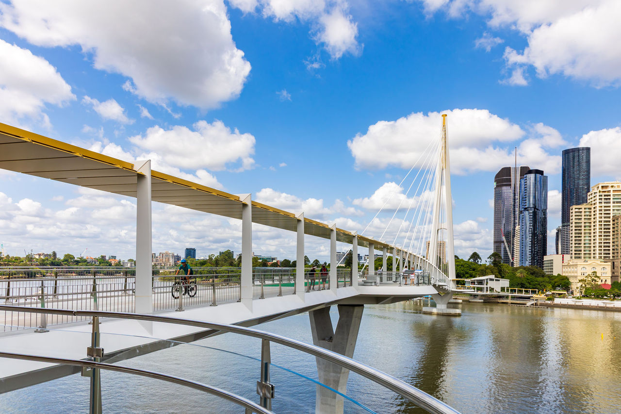 A cyclist rides across the Kangaroo Point Bridge. The bridge’s mast and city buildings can be seen in the background.
