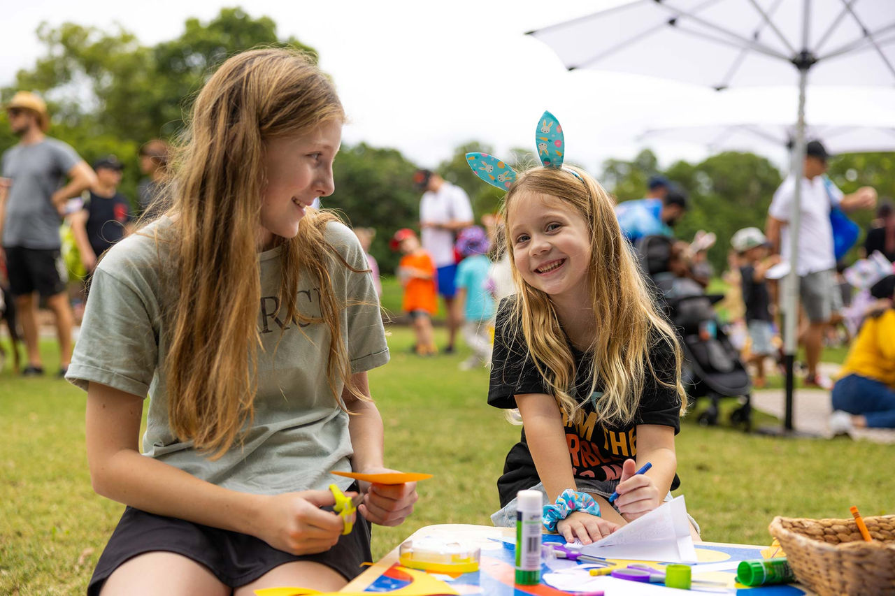 Two girls doing craft at a craft table at an Easter event. One is looking at the other and there are people in the background.