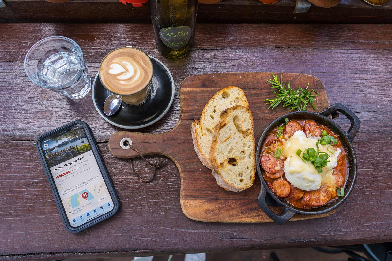 Brunch with cappuccino and water on a table with Brisbane app showing on a phone.