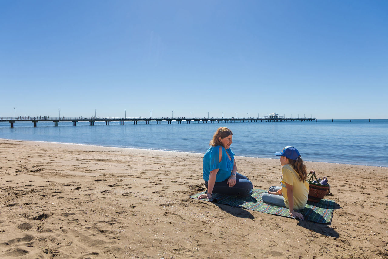 An adult and child sitting on a picnic blanket at Shorncliffe Foreshore. The pier is in the background.