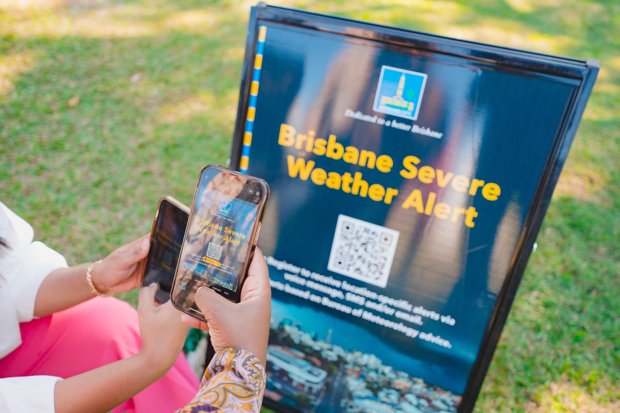 Close-up of two sets of hands scanning a QR code with smartphones on a Brisbane Severe Weather Alert A-frame sign at a festival.