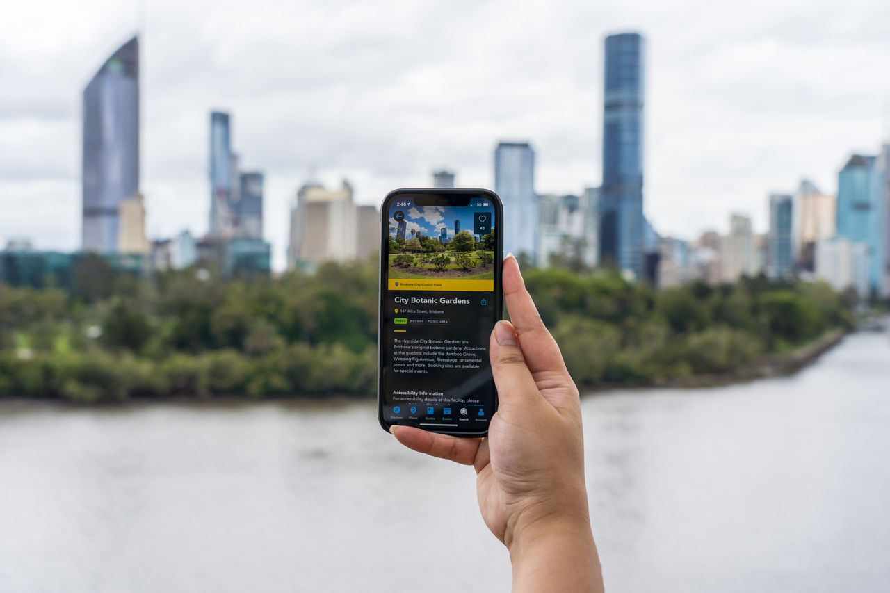 A hand holding a mobile phone showing the Brisbane app, looking over the Brisbane River.