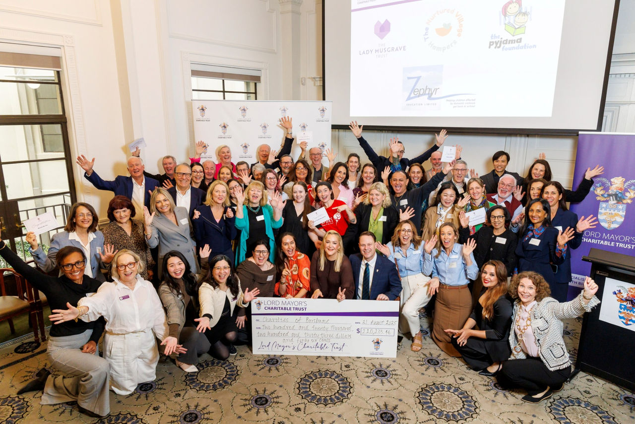 A large group of people smile with their hands up. At the front, a few people hold a large cheque with the “Lord Mayor’s Charitable Trust” written on it.
