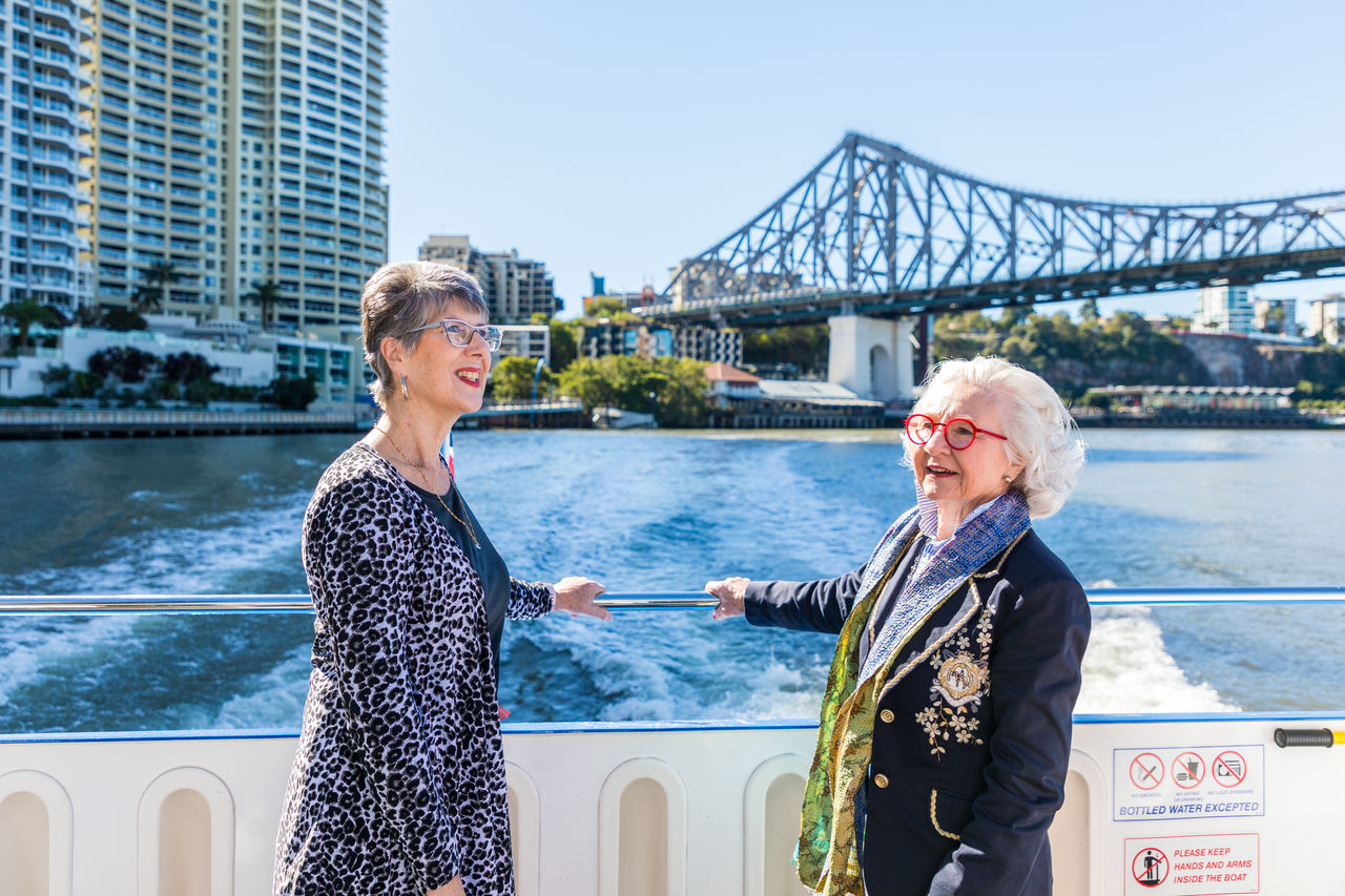 Two senior women travelling on a double-decker CityCat with Story Bridge in background.