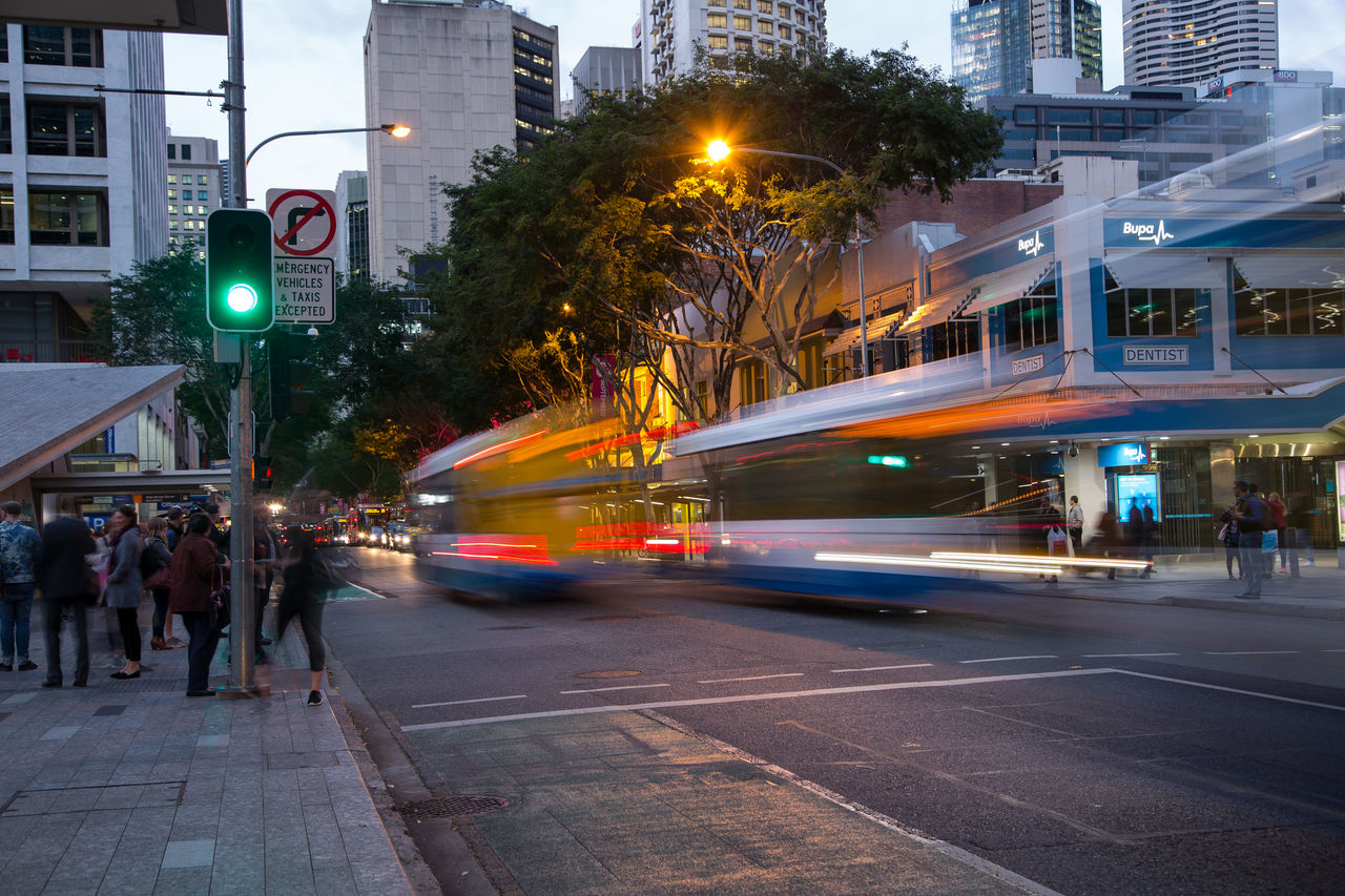 Traffic and pedestrians on Adelaide Street at dusk, including blurred images of 2 Council buses passing by King George Square.