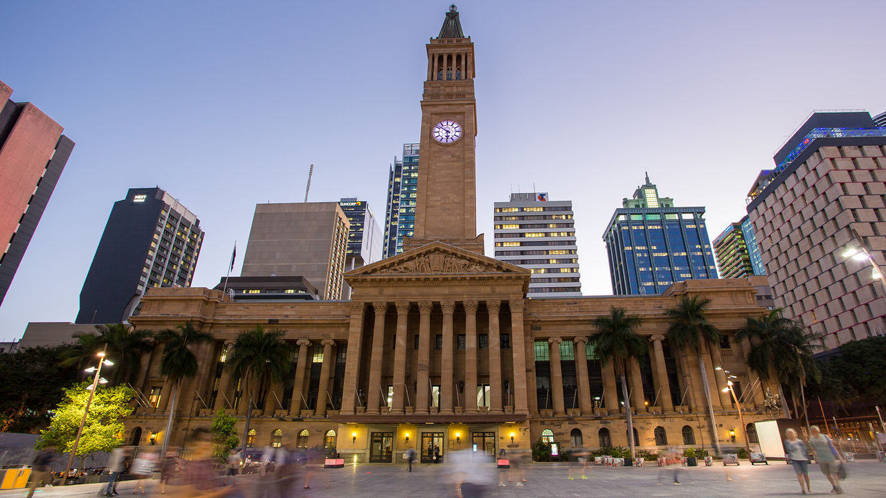 This image shows Brisbane City Hall at twilight. There are people walking past the building and buildings lit up in the background.