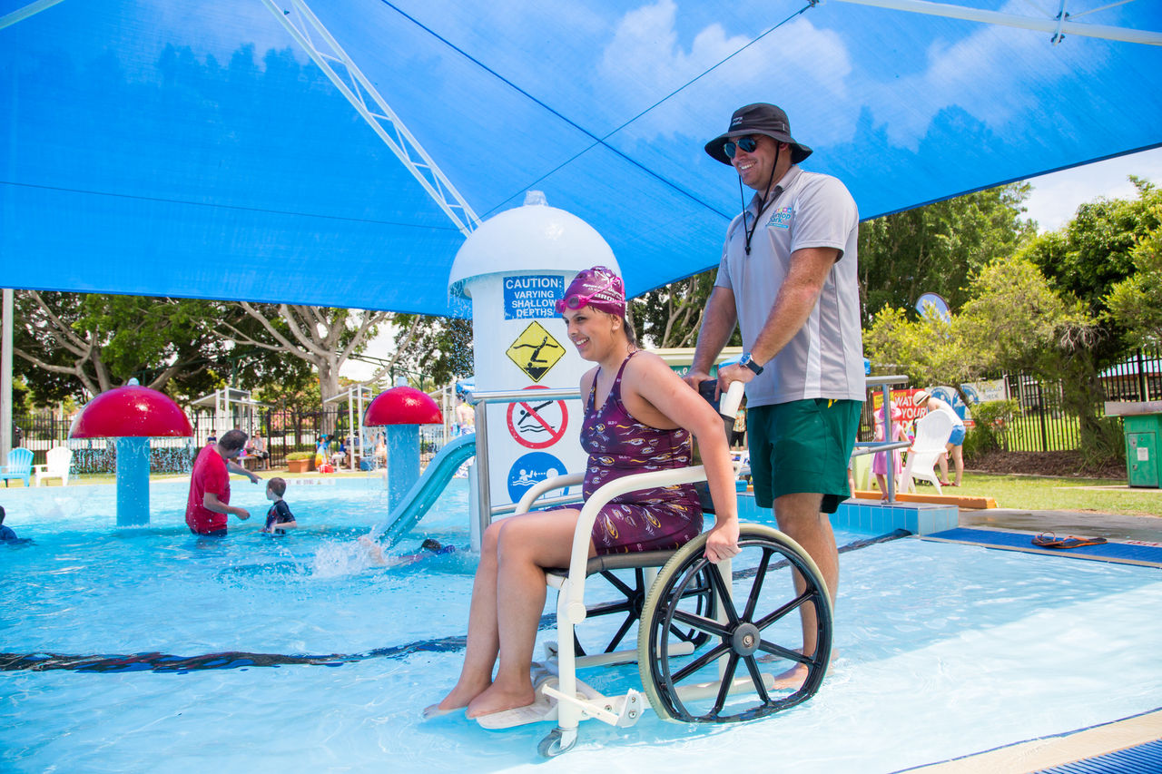 Woman in togs in a wheelchair on pool ramp with trainer at Dunlop Park Memorial Pool.